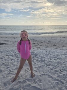  Girl in pink swimsuit and swim goggles standing on white sand beach with ocean in the background