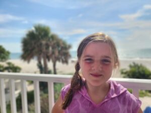 Girl with pink hoodie smiling on a beachside balcony with palm trees and ocean in background
