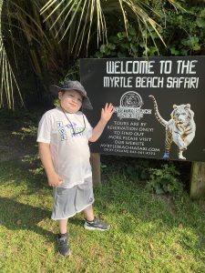 Nathan smiling in a Toby’s Dream Foundation shirt, standing next to the “Welcome to the Myrtle Beach Safari” sign.