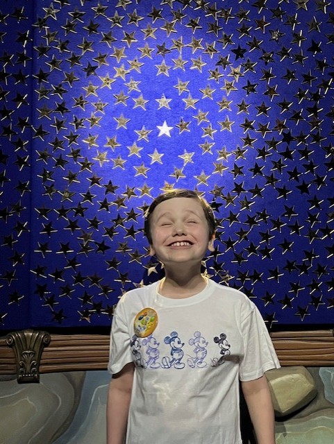 Mylo smiling joyfully in front of his star at Give Kids the World Village, surrounded by hundreds of golden stars representing children’s dreams come true.