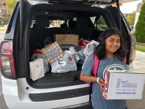Hailey smiles while holding a Toby’s Dream Foundation box beside a car filled with shopping bags from Macy’s, Old Navy, and Bath & Body Works during her dream shopping spree.