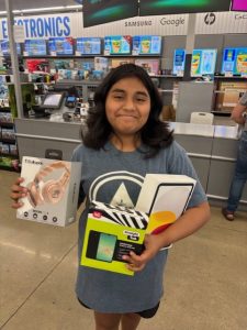 Hailey holds new headphones, a tablet, and a smartphone inside an electronics store during her Toby’s Dream Foundation shopping spree.