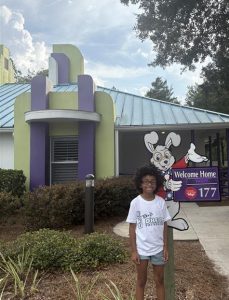 Girl smiling in front of Give Kids the World Village villa with Welcome Home sign.